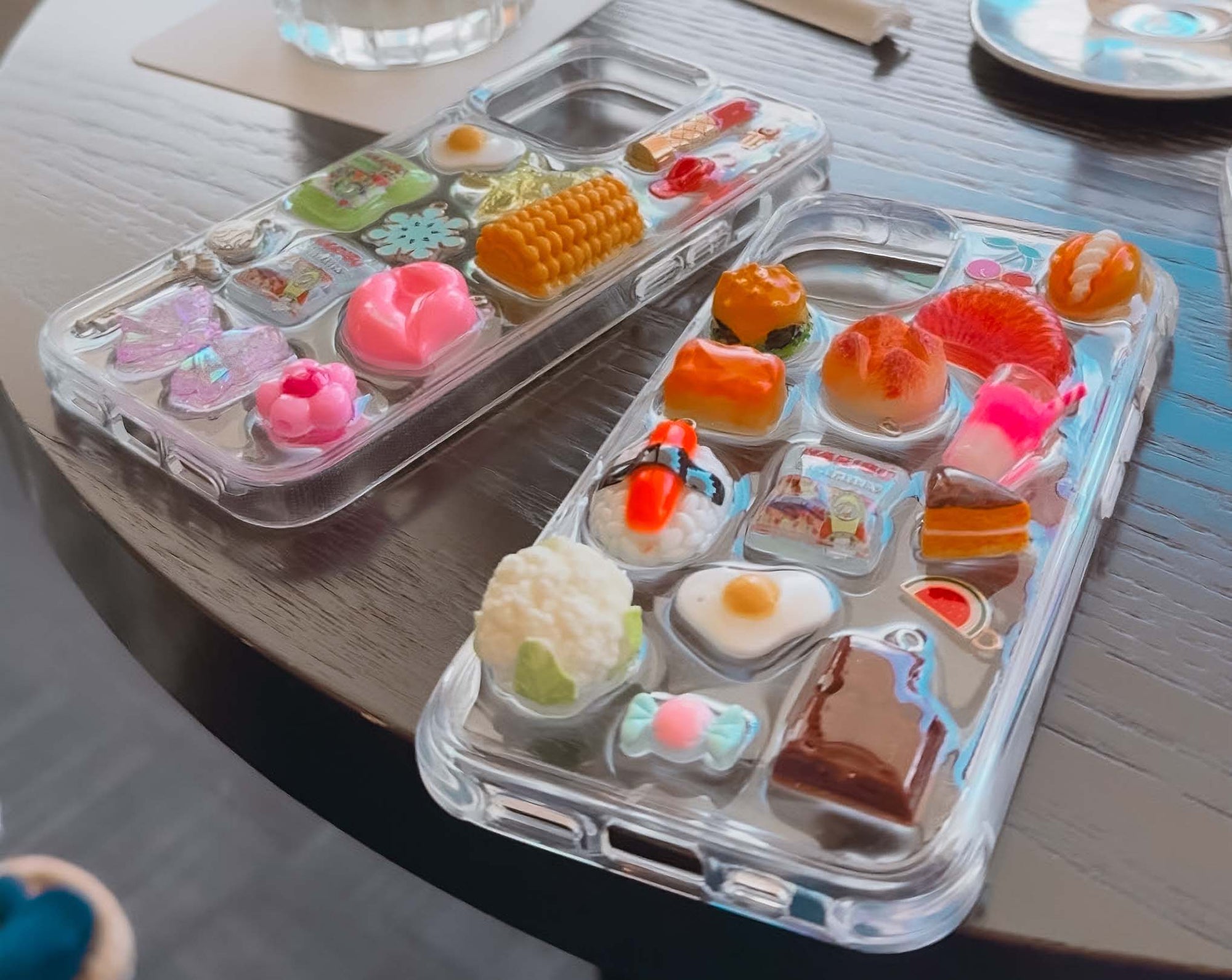 Two clear phone cases with chunky charms on a table.
