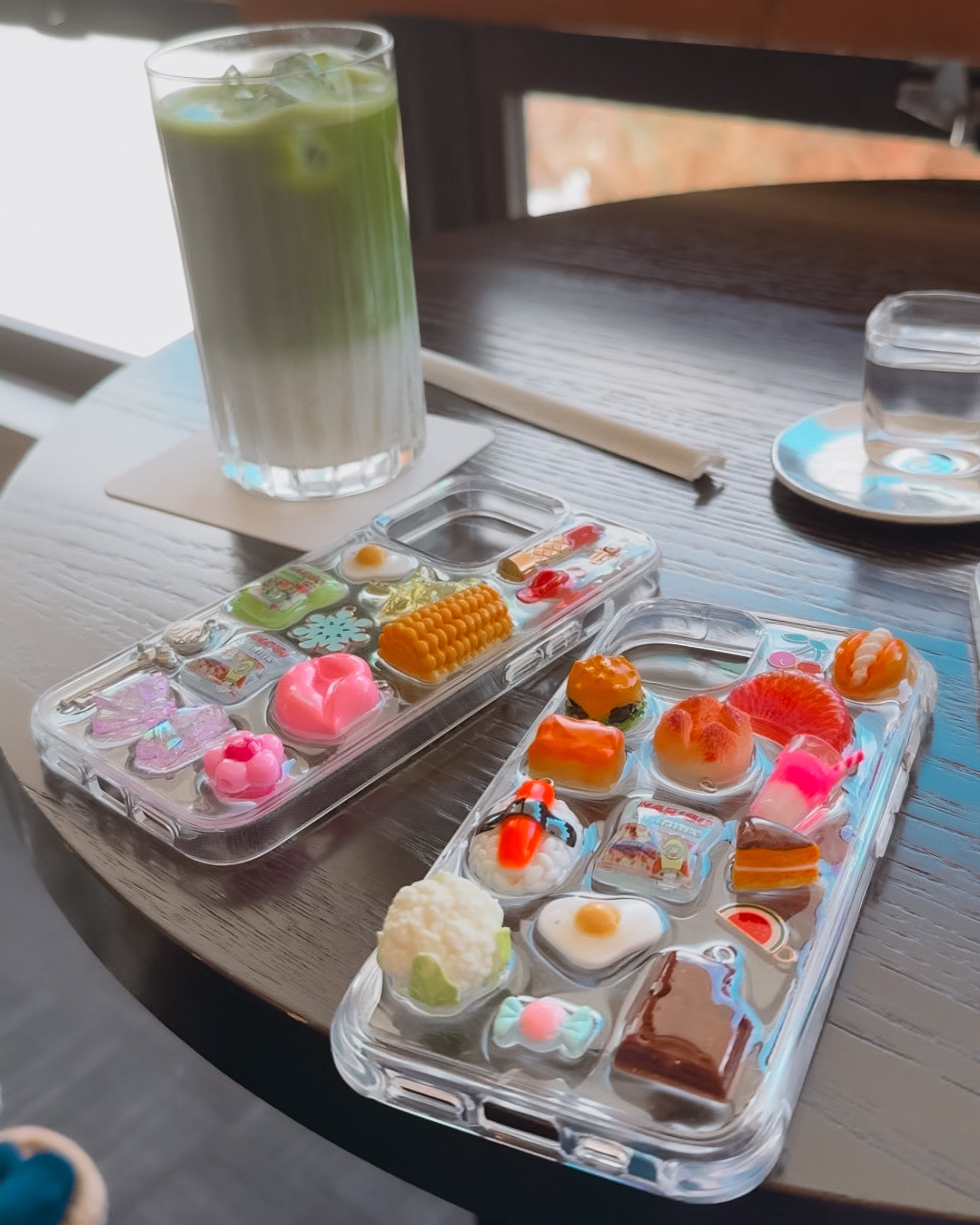 Two clear phone cases with chunky charms and a glass of iced matcha on a table.