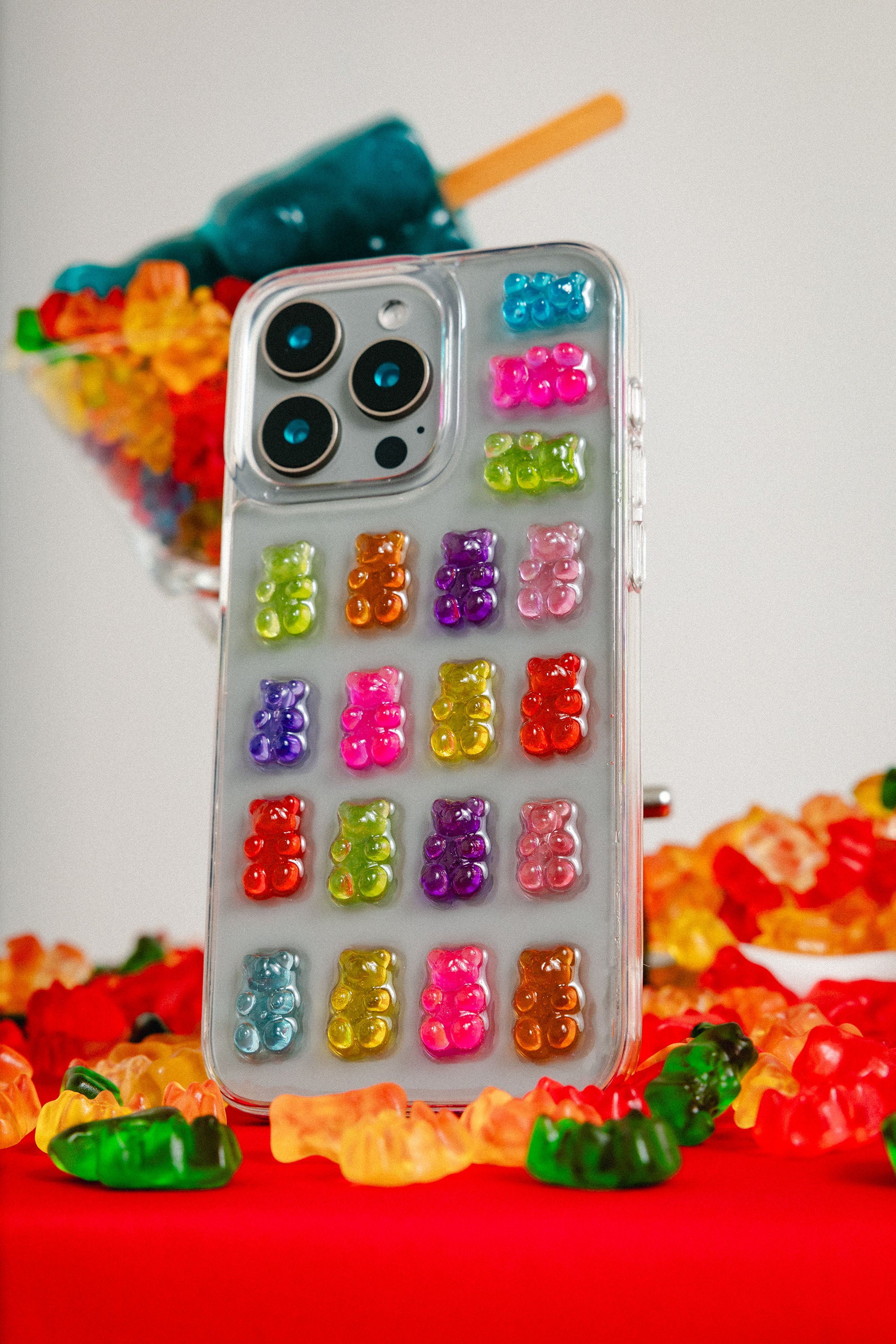 Phone case with gummy bear design surrounded by colorful candy on a red surface.