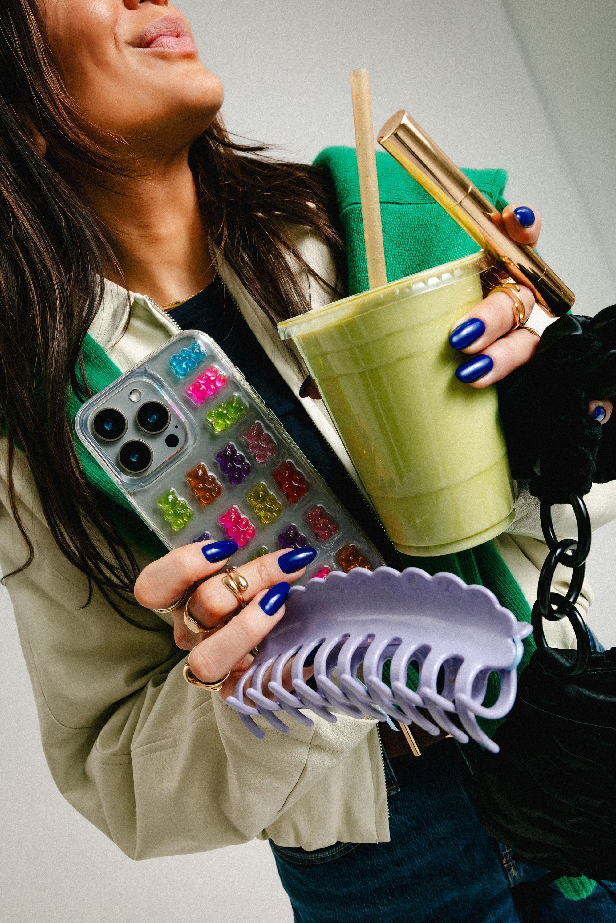 Woman holding a phone case with colorful gummy bear charms, a green cup with straw, and a purple hairclip.