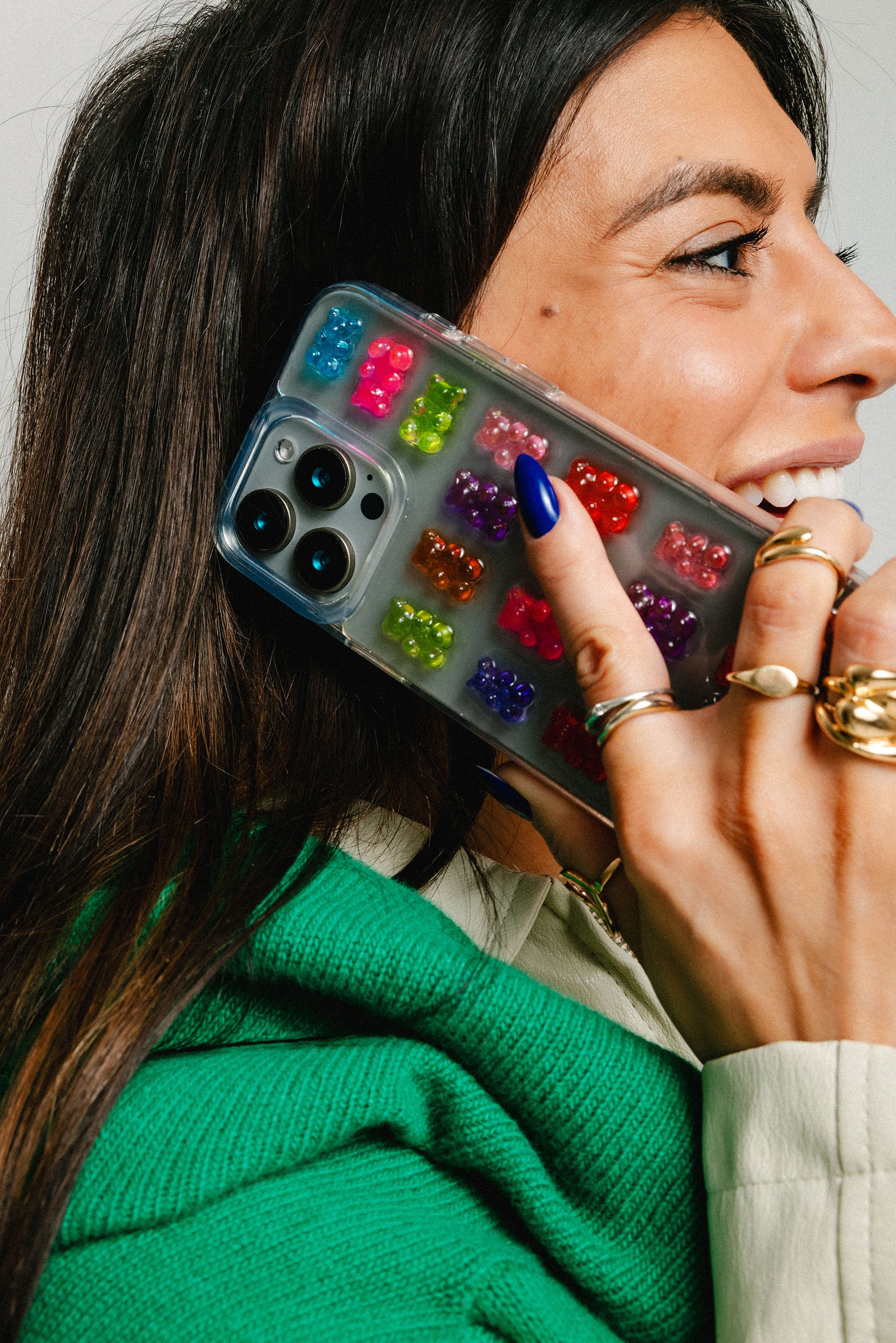 Woman holding a phone with a colorful gummy bear case, wearing a green sweater and gold rings.