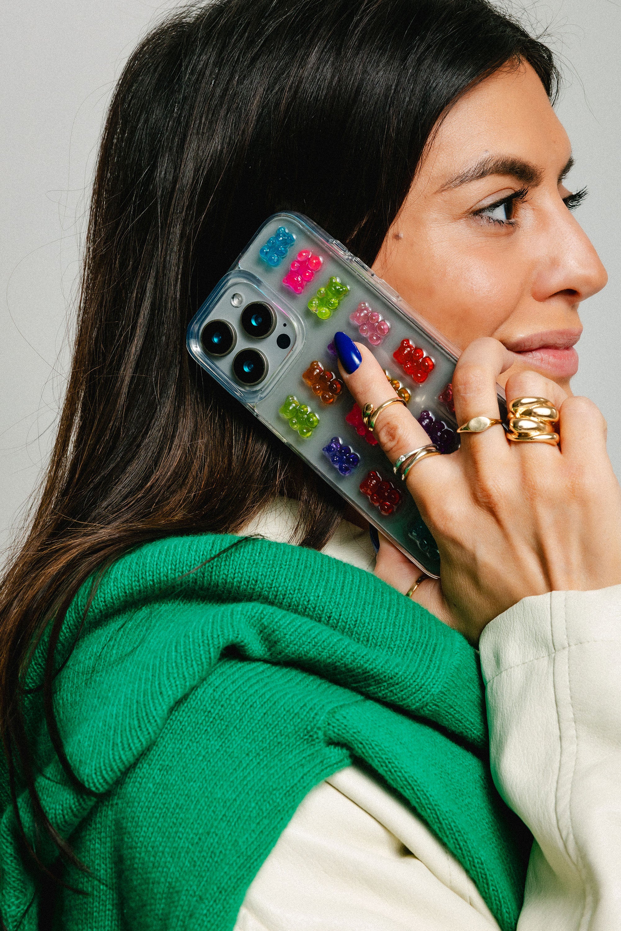 Woman wearing a green sweater and gold rings, holding a phone with a colorful gummy bear case against a neutral background